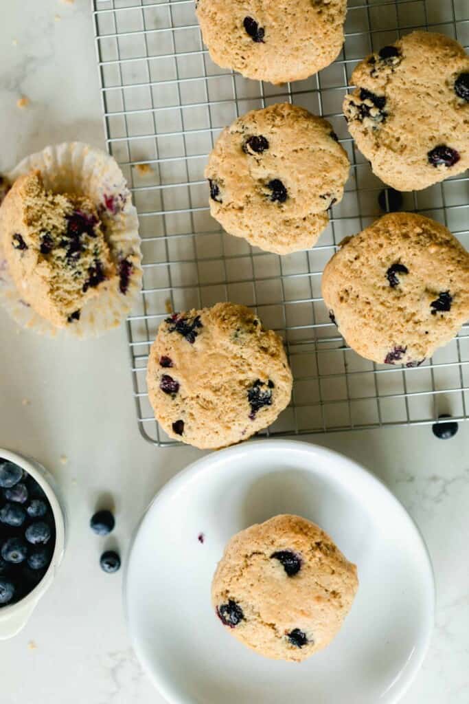 overhead photo of sourdough blueberry muffins on a wire rack with a muffin to the right cut in half and another muffin in the front on a white plate