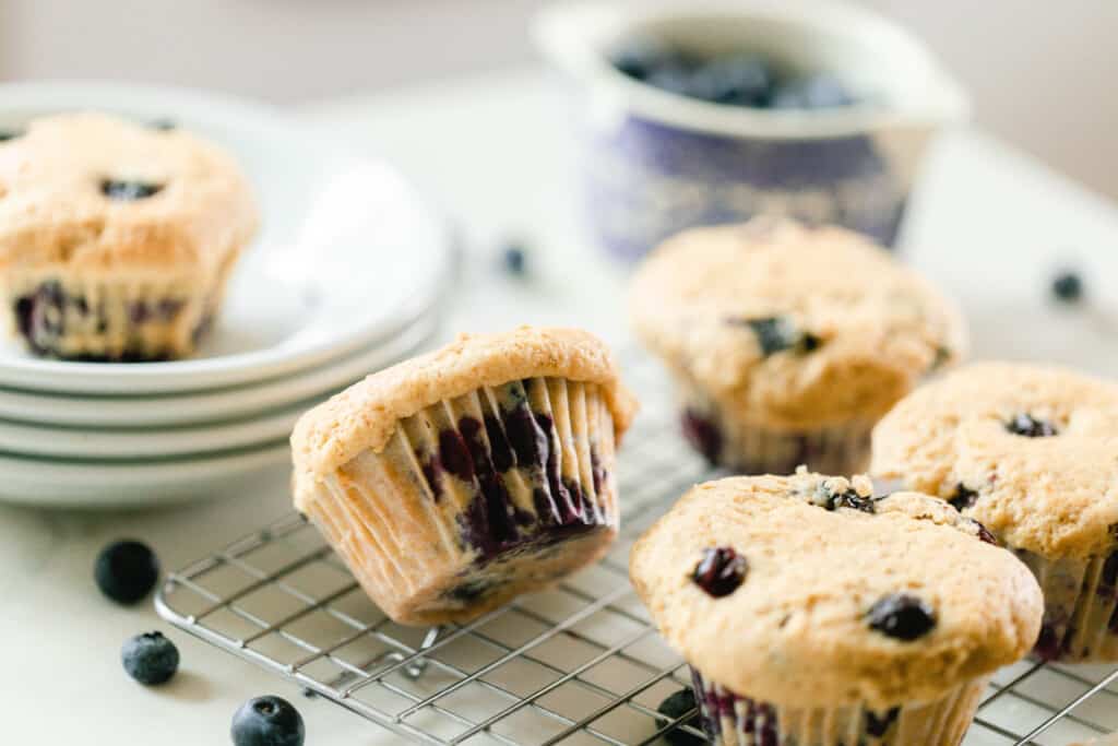 sourdough blueberry muffins cooling on a wire rack on a white countertop with 4 white plates stacked with a muffin on top to the left and a bowl of blueberries in the background