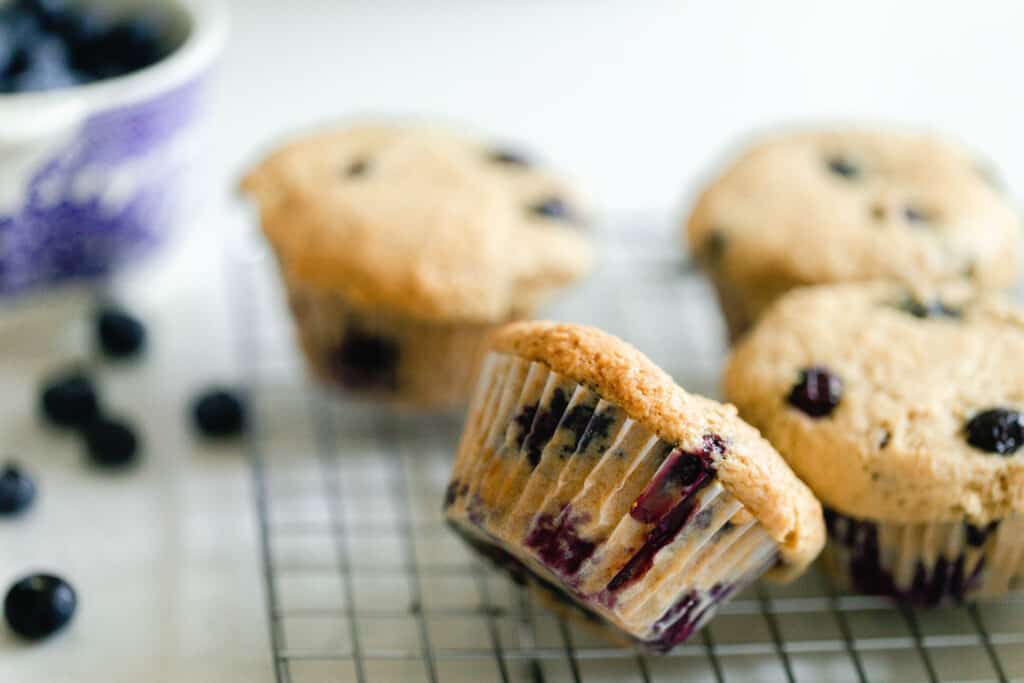four sourdough blueberry muffin on a wire rack with one of the muffins tipped over and leaning on another muffin. Blueberries are scattered on the countertop and a small bowl to the left