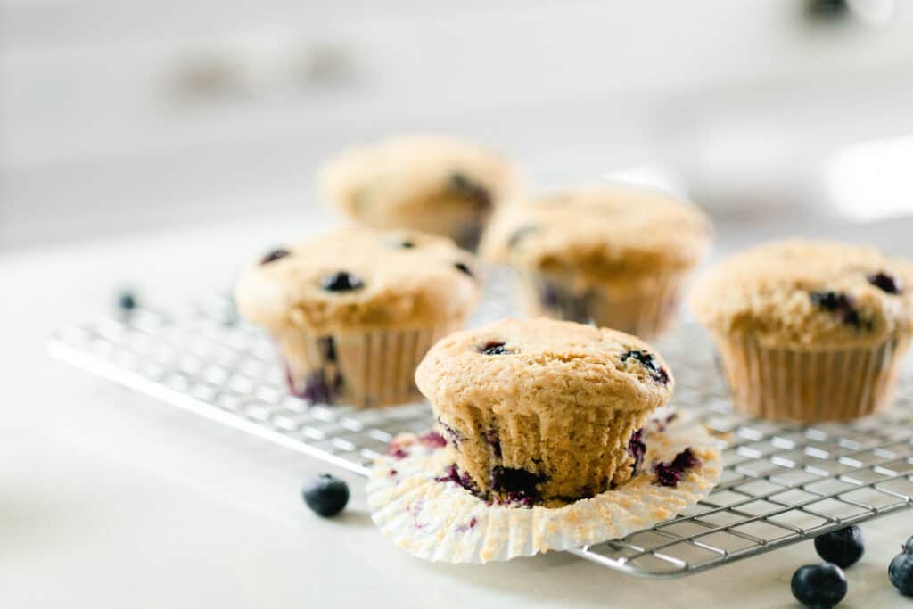 five sourdough discard blueberry muffins on a wire cooling rack on a white countertop with fresh blueberries sprinkled around. The muffin at the corner has the cupcake liner peeled down