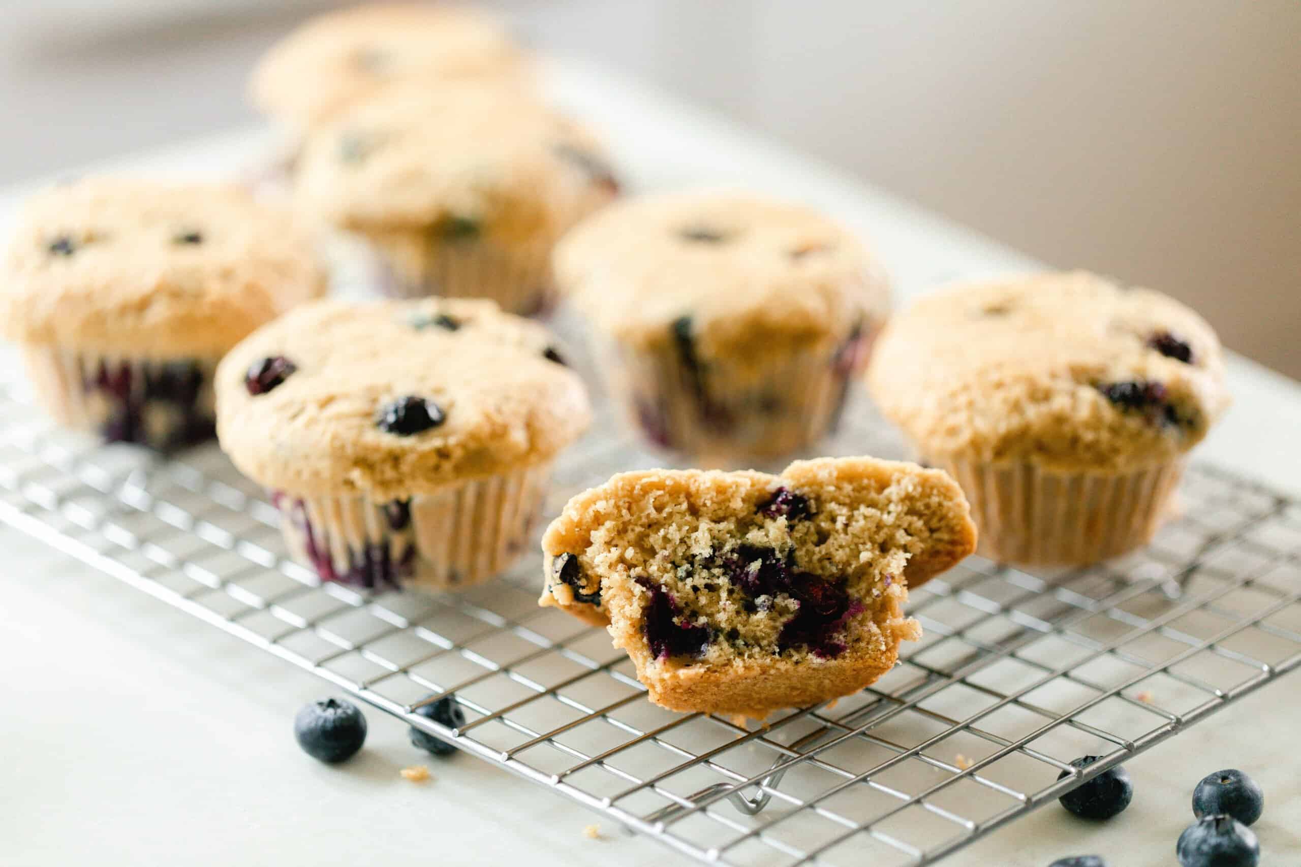 sourdough blueberry muffins on a wire rack on a white countertop with blueberries scattered around on the counter. The front muffin is sliced in half.