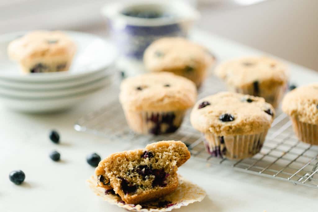 sourdough blueberry muffin sliced in half on a white countertop with blueberries to the left. A wire rack with 5 more muffins and four white plates stacked up with a muffin on top