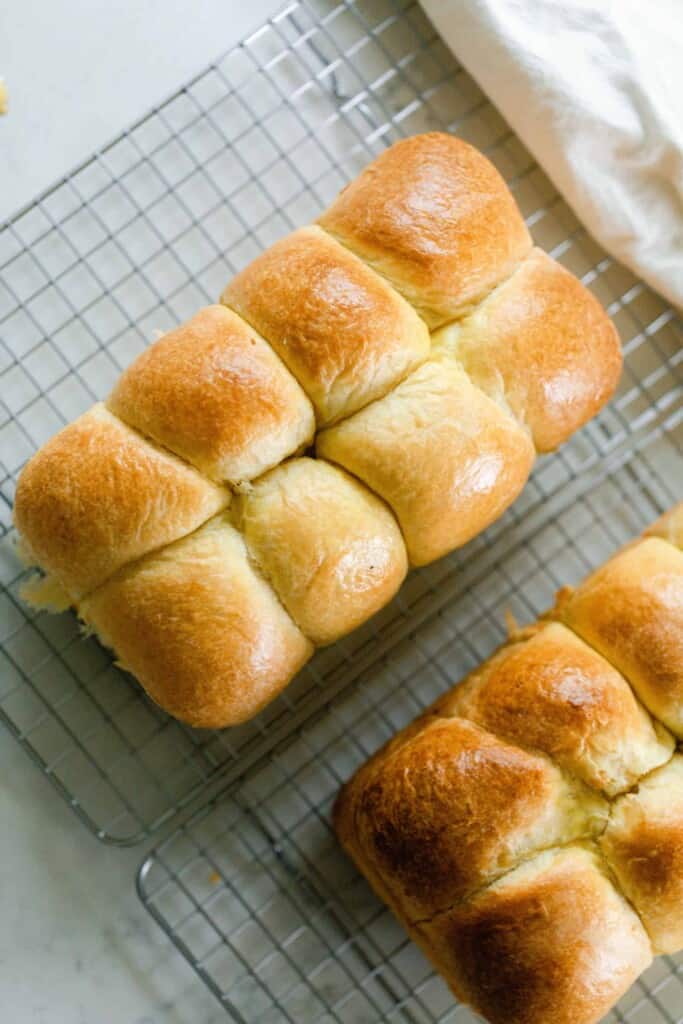 overhead photo of two loaves of sourdough brioche with a golden crust on a wire rack on a white countertop with a white towel in the back right corner