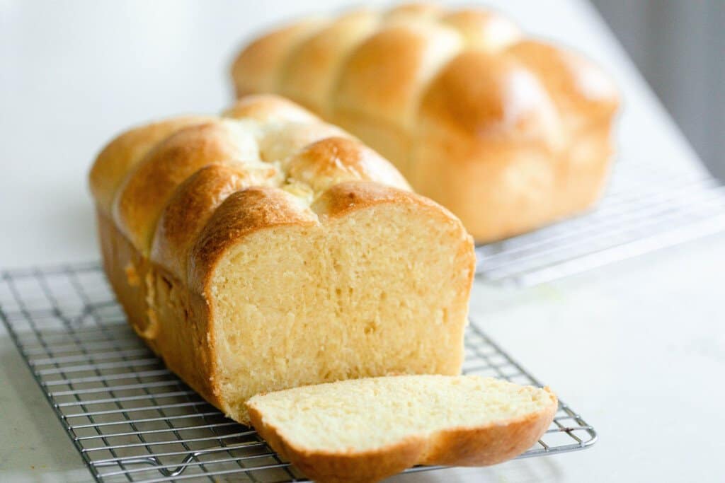 a loaf of sourdough brioche on a wire rack with a slice cut out. Another loaf is in the background