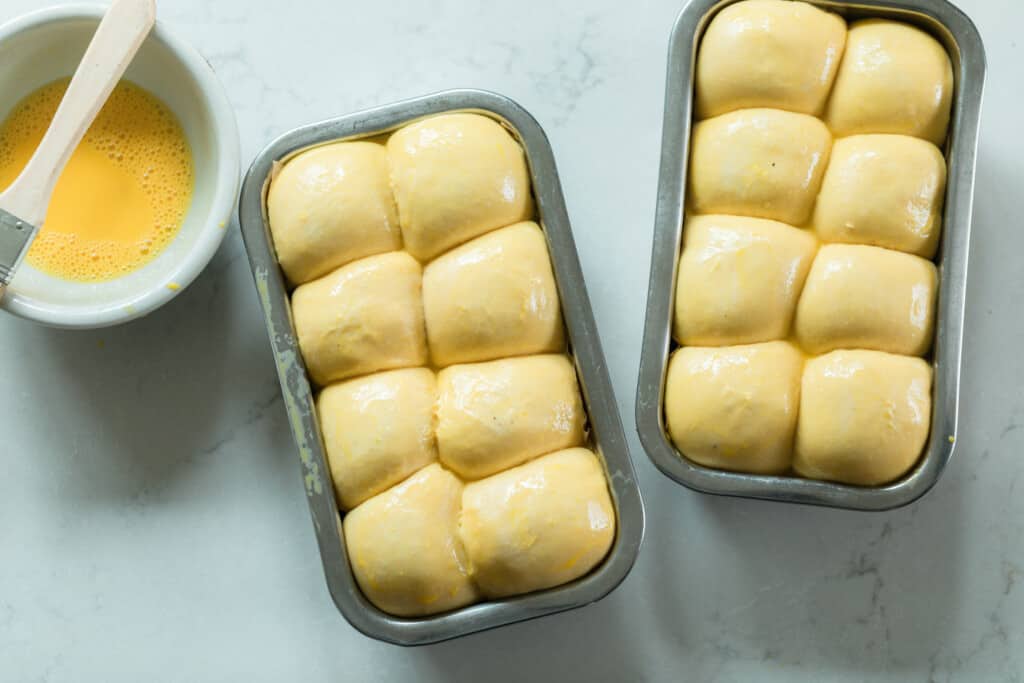 two stainless loaf pans with sourdough brioche dough that has risen and glazed with an egg wash on a white countertop. A white bowl with an egg wash and a pastry brush resting on the rim of the bowl sit to the let of the loaves.