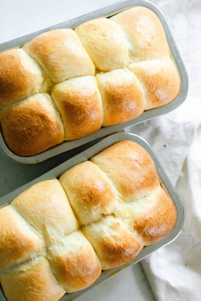 overhead photo of two loaves of sourdough brioche in stainless loaf pans on a white countertop and white towel