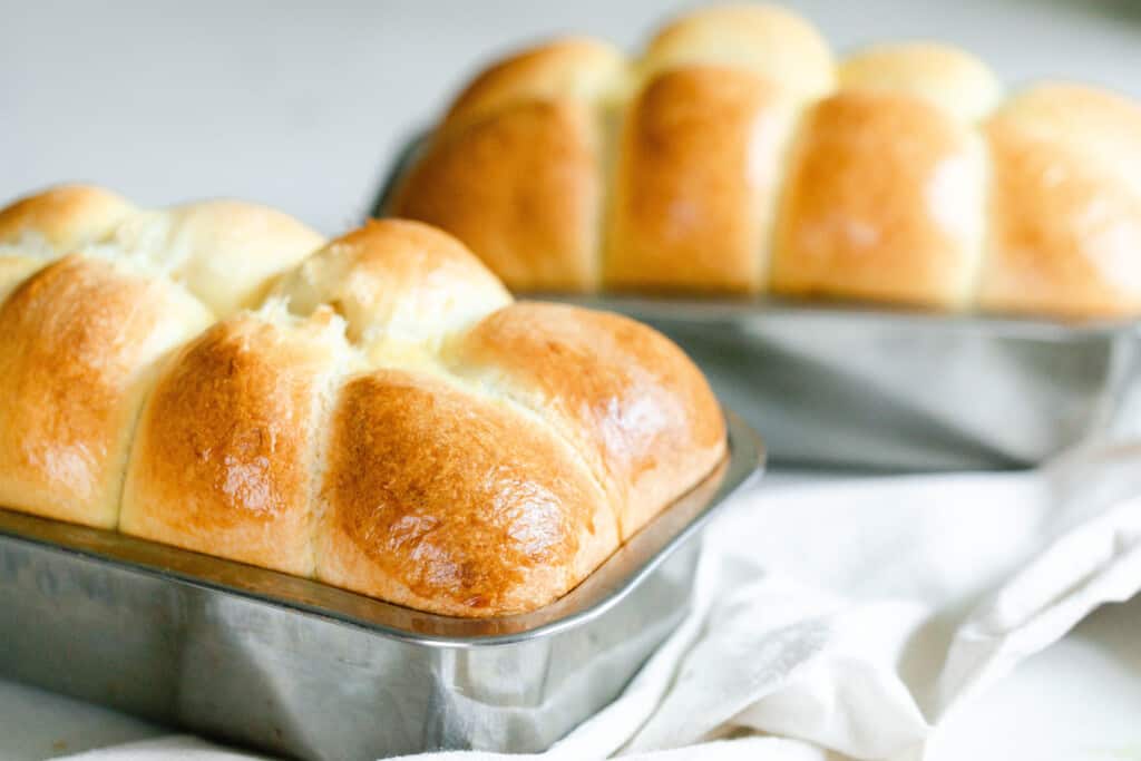 close up photo of sourdough brioche in a stainless steel loaf pan on a white towel with another loaf in the background