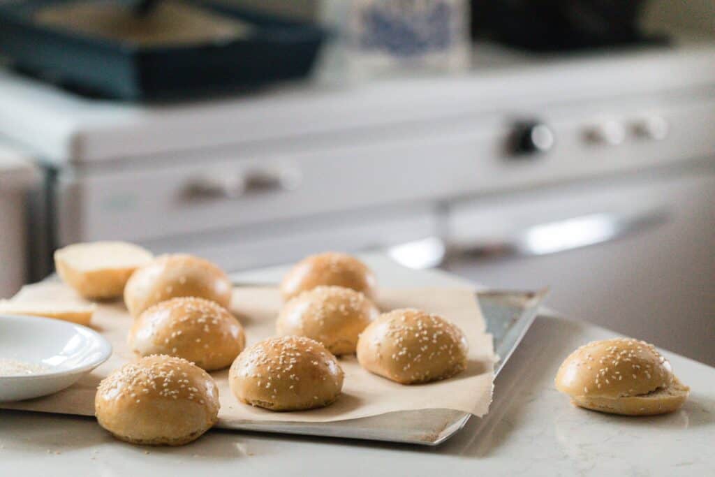 fluffy sourdough hamburger buns scattered on a parchment lined baking sheet and one on a white countertop on a kitchen island. A vintage stove is in the background