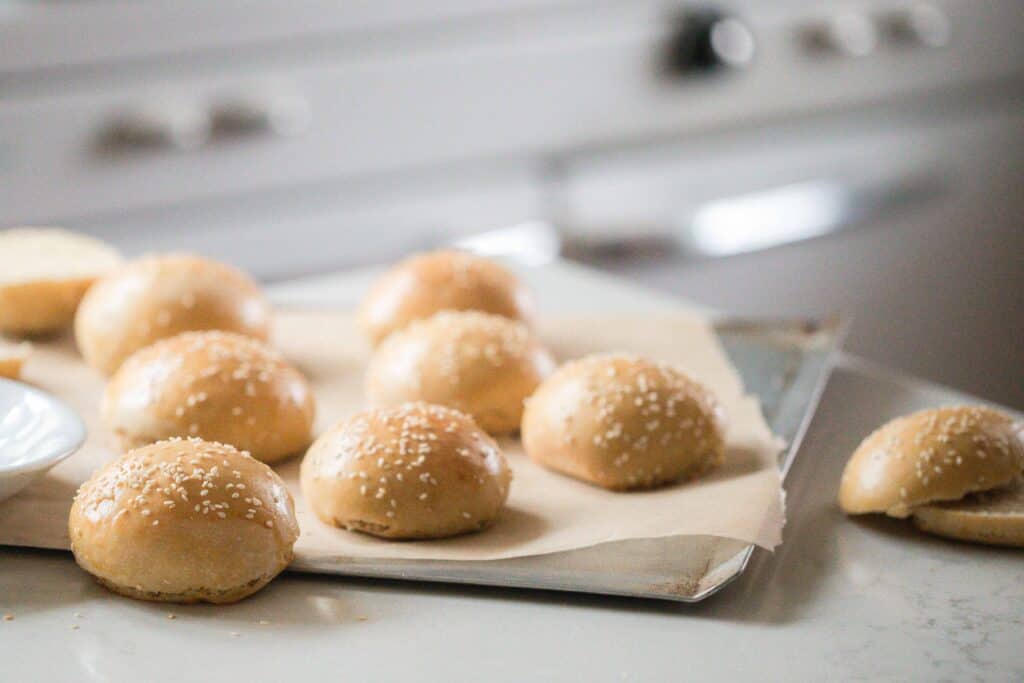 Sourdough buns topped with sesame seeds on a parchment lined baking Sheed on a white quartz countertop