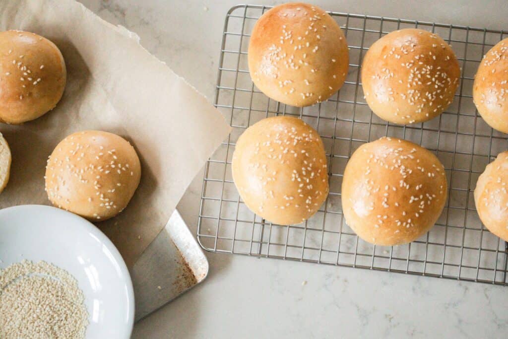Overhead photos of sourdough buns on a wire baking rack  on a quartz countertop with more buns to the left on a baking sheet