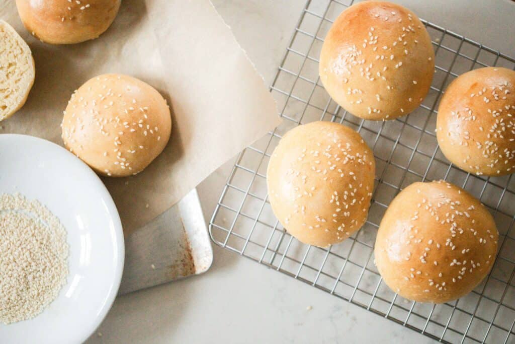 Overhead photo of sourdough hamburger buns on a wire rack with a sheet pan to the left of hamburger buns on parchment line baking sheet with a bowl of sesame  seeds.
