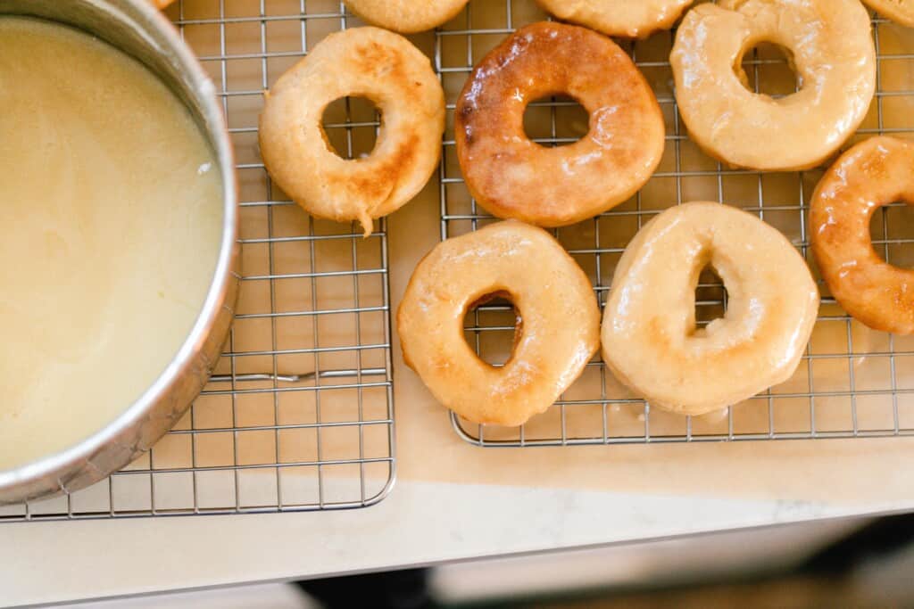sourdough donuts on a wire rack with parchment paper underneath with a pot of donut glaze to the left