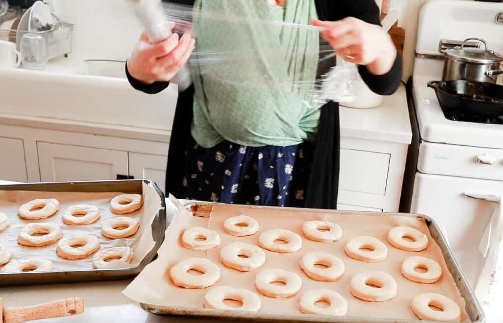 woman wearing a baby is covering sourdough donut dough with plastic wrap to rise.