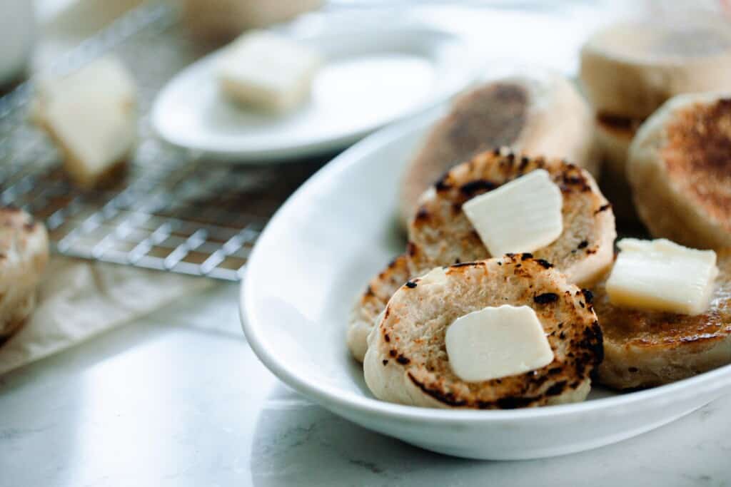 sliced sourdough English muffins with  pats of butter on top in a white bowl on white countertop  with other kitchen things in the background