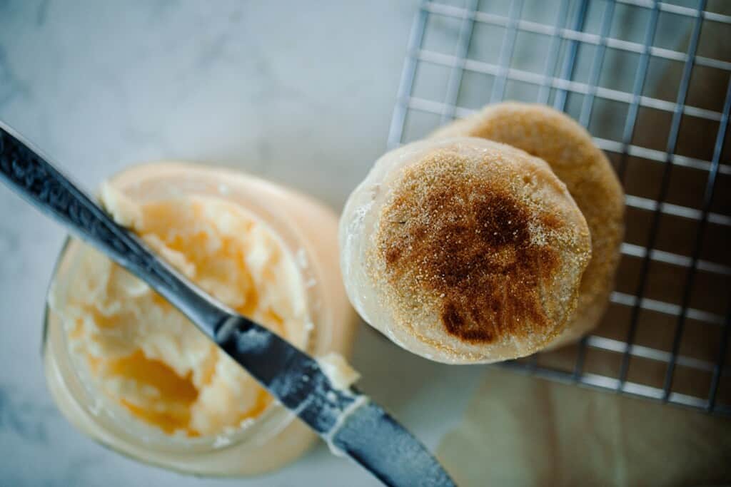 overhead photos of two sourdough English muffins stacked up on a wire cooling rack with a small mason jar of butter with a knife resting on top to the left