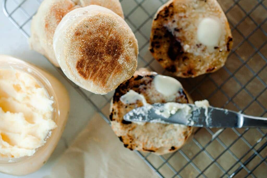 knife buttering a sourdough English muffin half on a wire cooling rack over parchment paper. Two more muffins sit directly behind and a jar of butter to the left