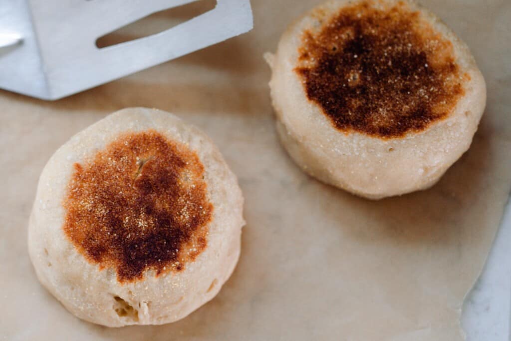 two sourdough English muffins on parchment paper with a metal spatula behind.