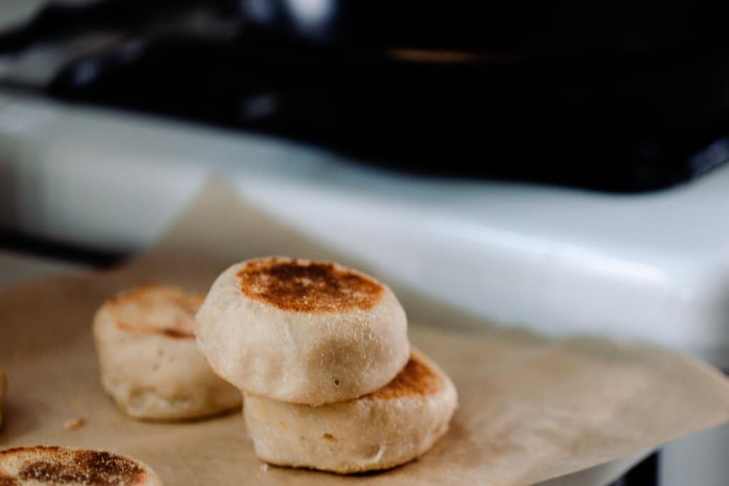 three sourdough English muffins stacked on parchment paper with a white antique stove in the background