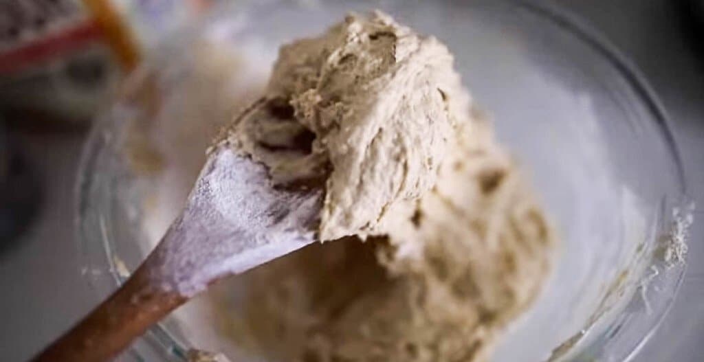 a wooden spoon with a scoop full of sourdough English muffin dough. The spoon is held above a glass bowl of remaining dough