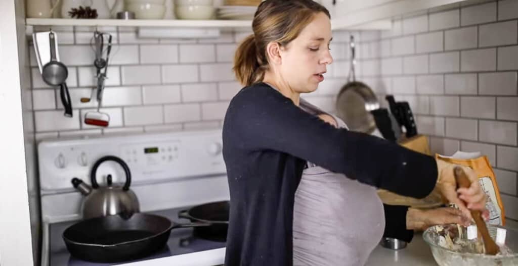 woman wearing a baby is in her kitchen staring dough together in a glass bowl with a wooden spoon.