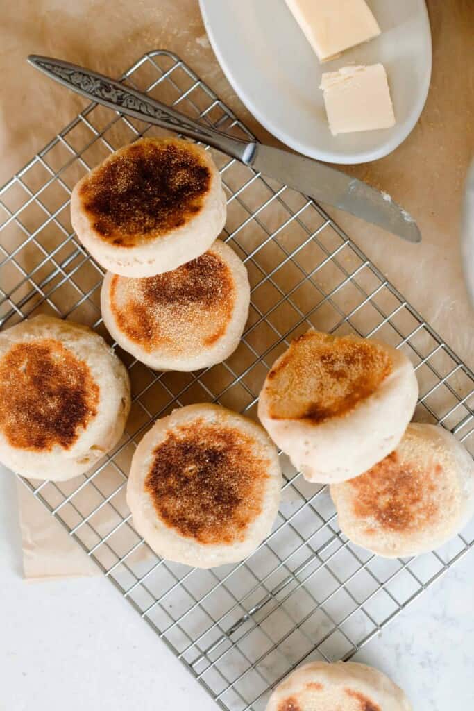 seven sourdough English muffins spread out on a wire cooling rack over parchment paper with a butter knife resting on the right corner. A small white dish with a stick of butter is to the right