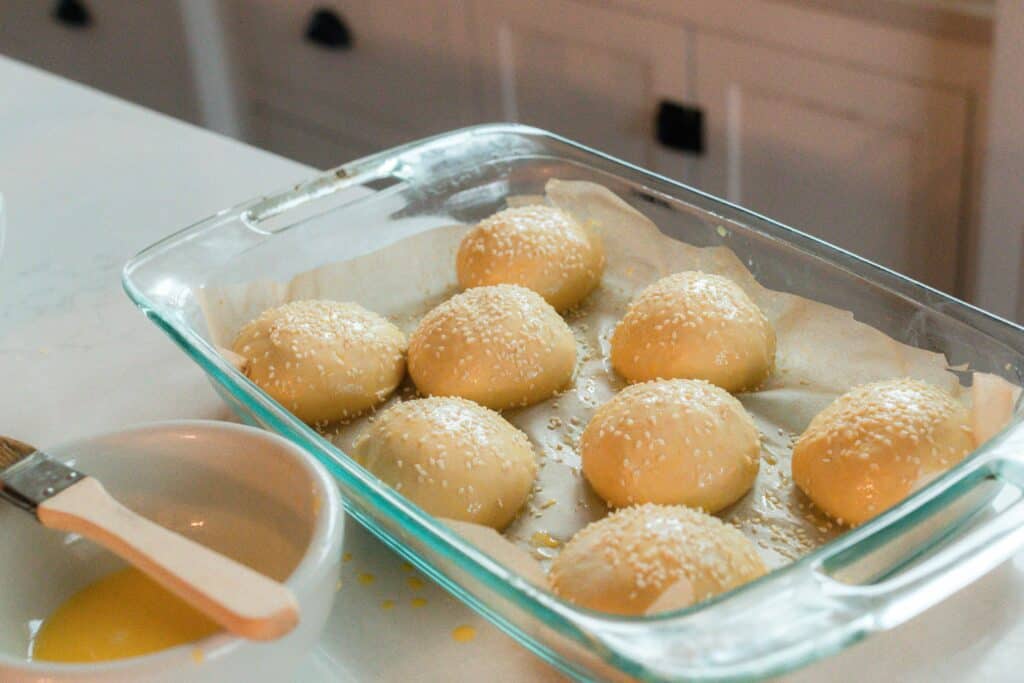 Sourdough hamburger bun dough on a parchment lined glass baking sheet with an egg wash and sprinkled with sesame seeds.