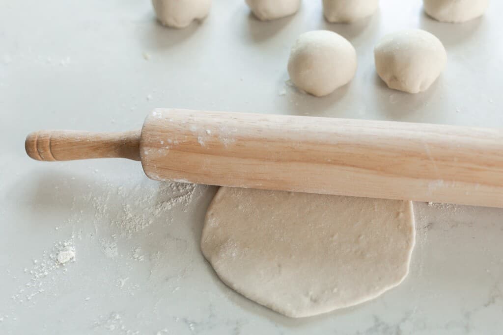 sourdough tortilla dough being rolled out with a rolling pin, balls of dough are behind the rolling pin