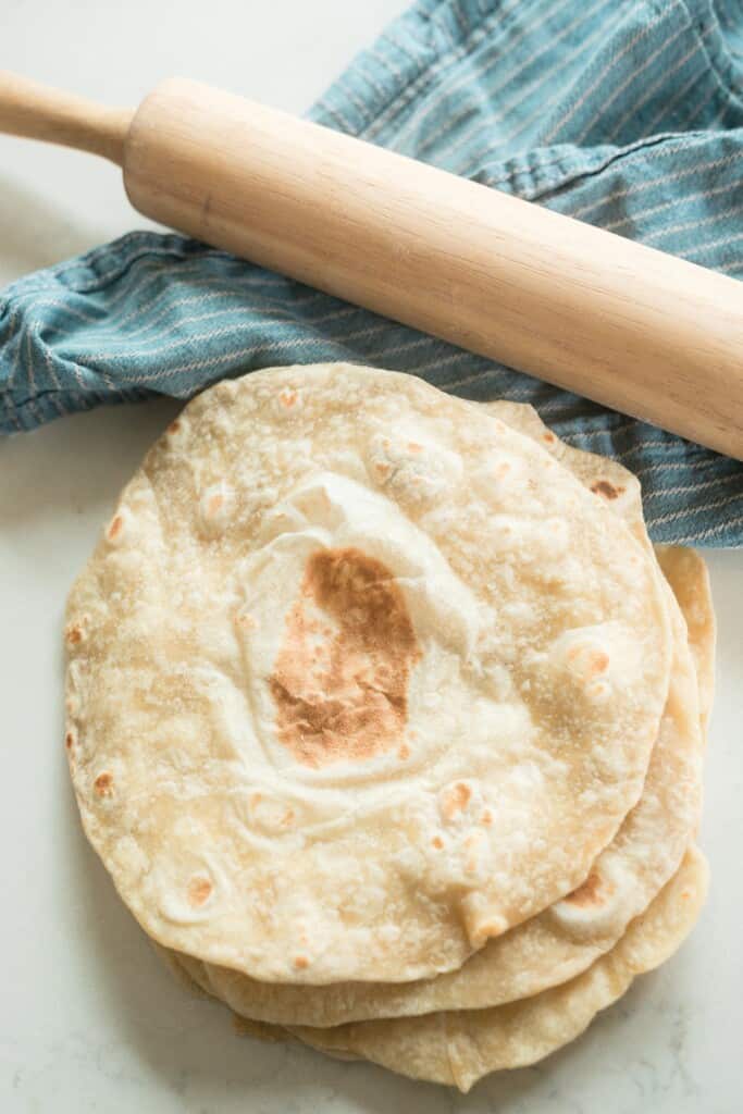 overhead photo of sourdough tortillas stack in a pile on a white quartz countertop with a blue stripped towel in the background with a rolling pin on top