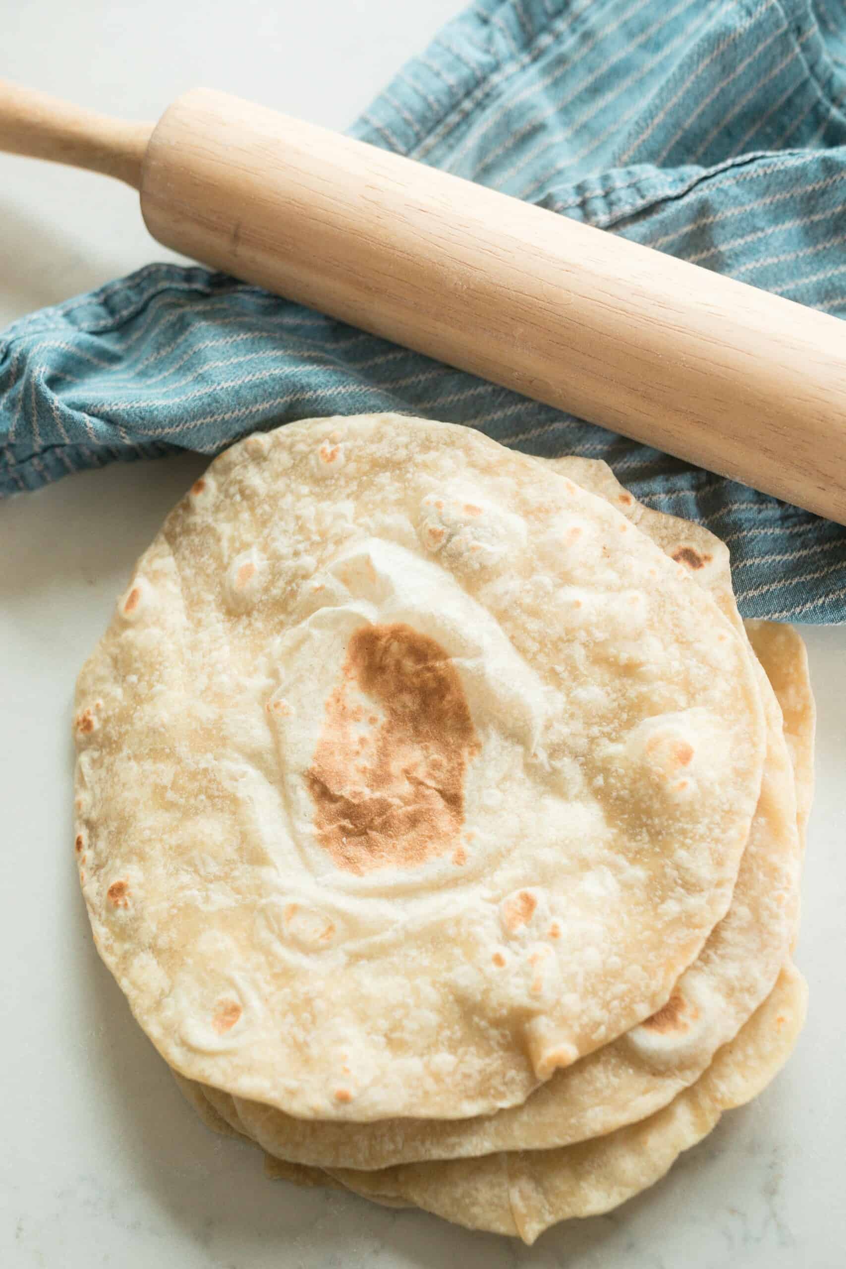 overhead photo of sourdough tortillas stack in a pile on a white quartz countertop with a blue stripped towel in the background with a rolling pin on top