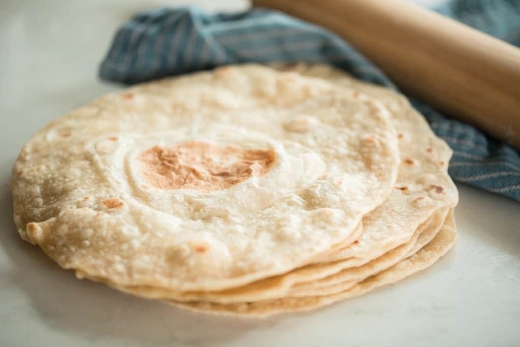 sourdough tortillas stacked up on a white quartz countertop with a folded blue towel with a rolling pin in the back right corner