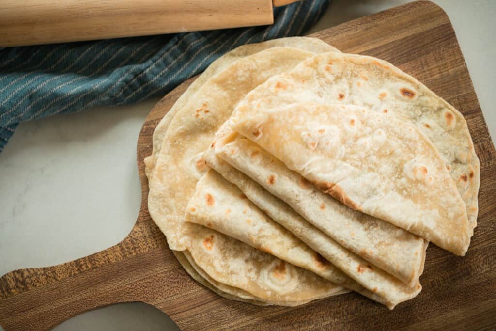 overhead photo of sourdough tortillas folded in half on a wood cutting board with a blue towel with a rolling pin to the back left