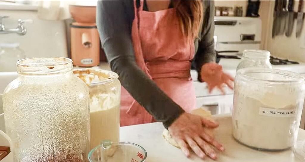 woman wearing a pink apron kneading sourdough English muffin dough on a white countertop surrounded by other muffin ingredients