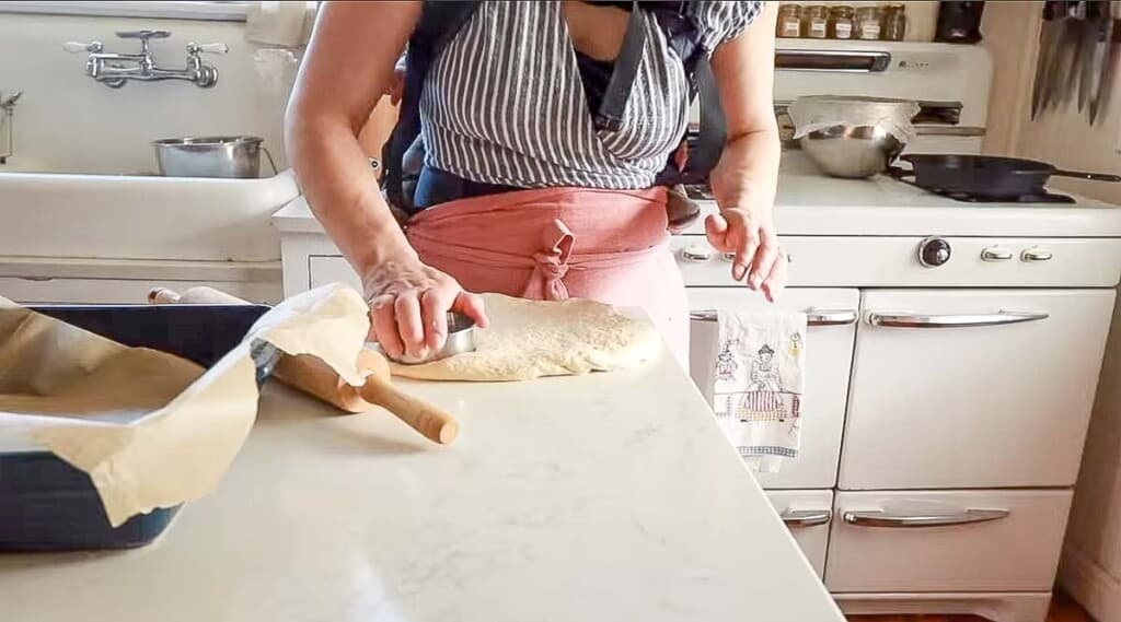 Woman in a pink apron cutting out sourdough English muffins with a biscuit cutter on a white quartz countertop