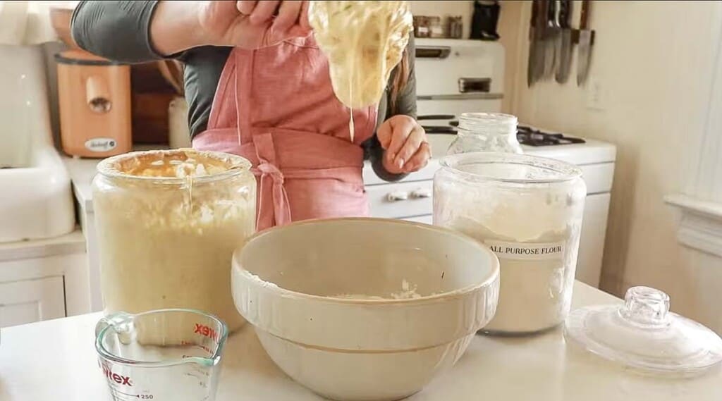 a woman in her white farmhouse kitchen adding active sourdough starter to a large bowl with flour
