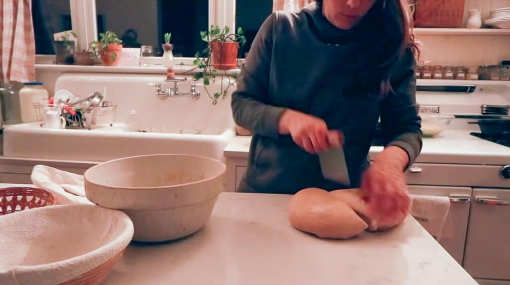 woman in a gray sweatshirt cutting sourdough bread dough in half with a bench scraper on a white countertop 