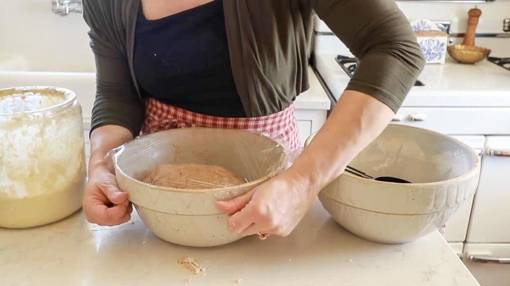 woman placing plastic wrap over whole wheat sourdough bread dough in a stoneware bowl in a white kitchen 