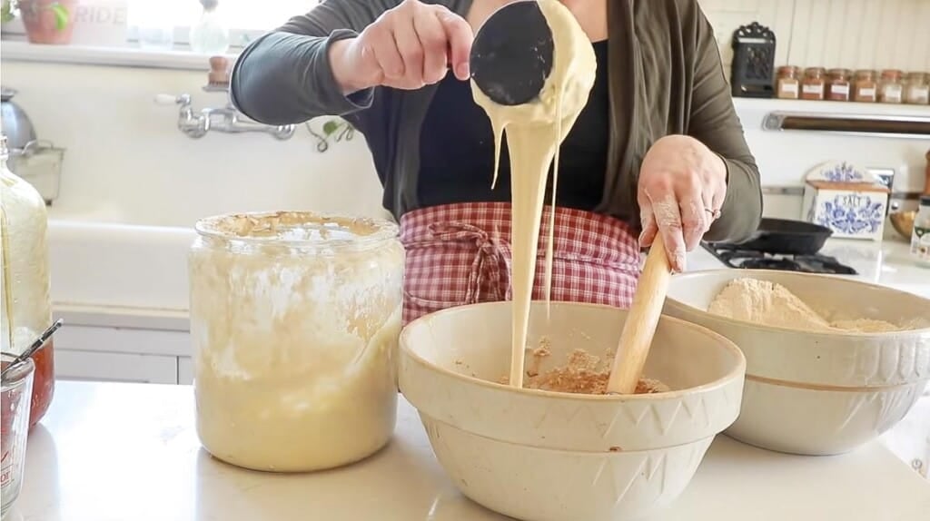 woman wearing a black shirt and gray sweater adding sourdough starter to a large stoneware bowl