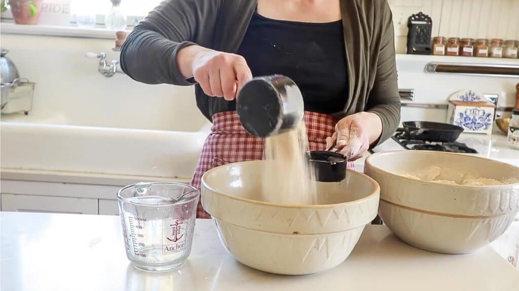 woman standing in her white kitchen adding freshly ground flour into a stone wheat bowl