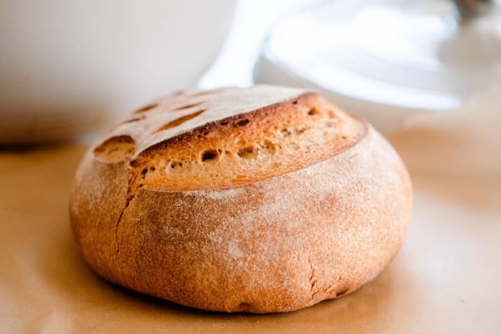 side view of a loaf of whole wheat sourdough bread boule on parchment paper with a white bowl in the background