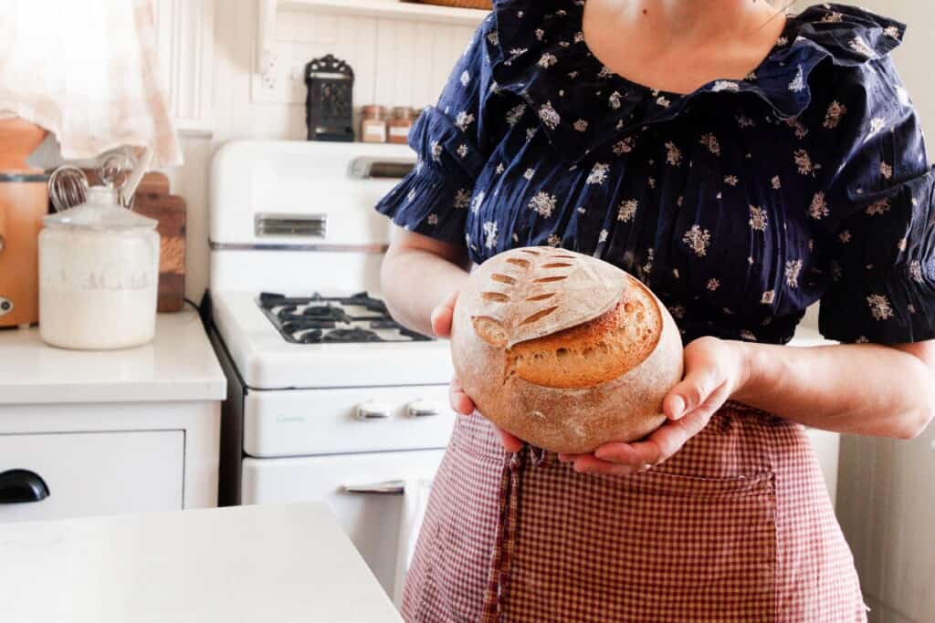 woman wearing a blue dress and wearing a red apron holding a whole wheat sourdough bread boule. Standing in a white kitchen