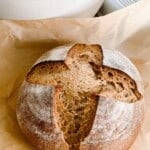 whole wheat sourdough bread boule with a cross pattern on parchment paper with a white dutch oven in the background