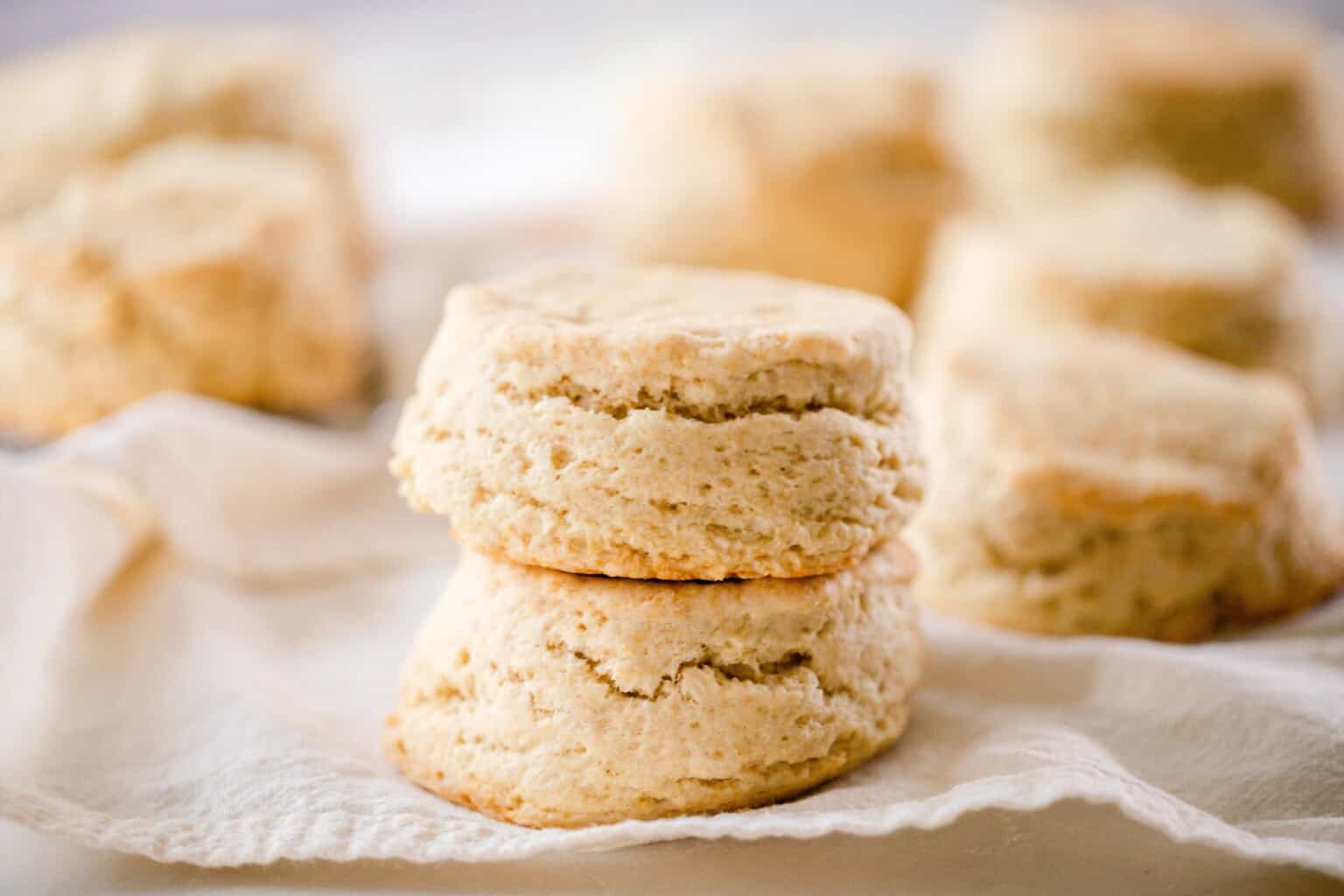 Two fluffy sourdough biscuits stacked on top each other on a linen towel with stacks of more biscuits in the background.