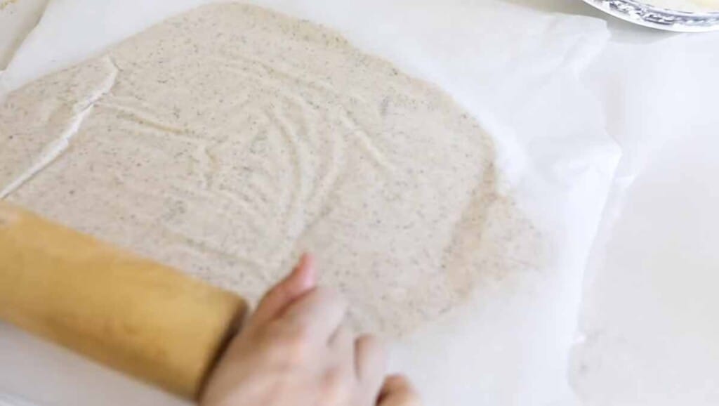 hands holding a rolling pin rolling out sourdough cracker dough between two pieces of parchment paper