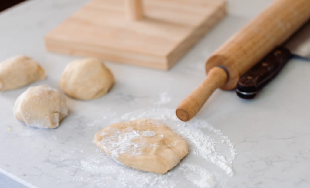 sourdough pasta dough patted out on a white countertop with more dough balls to the left, and a rolling pin and knife to the top right
