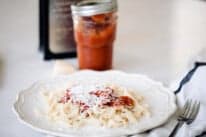freshly cooked sourdough pasta topped with homemade marinara and grated parmesan cheese on a white fluted plate with a white and blue towel with a fork placed on top to the right. A jar of marina, cheese grater and a chunk of parmesan cheese are behind the plate.