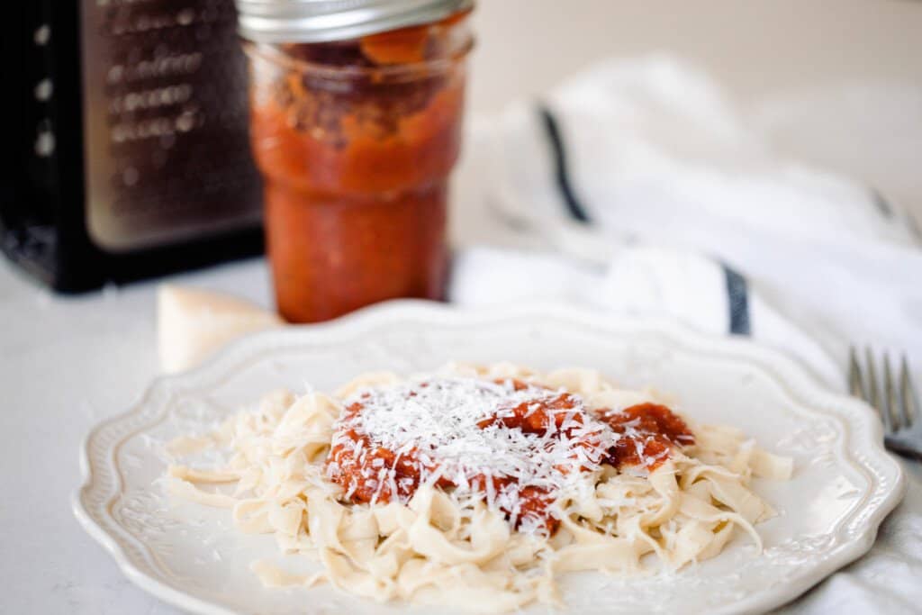 homemade sourdough pasta topped with marinara sauce and shredded cheese on a white fluted plate. A white towel with a blue stripe, jar of marinara sauce, and cheese grater is in the background.