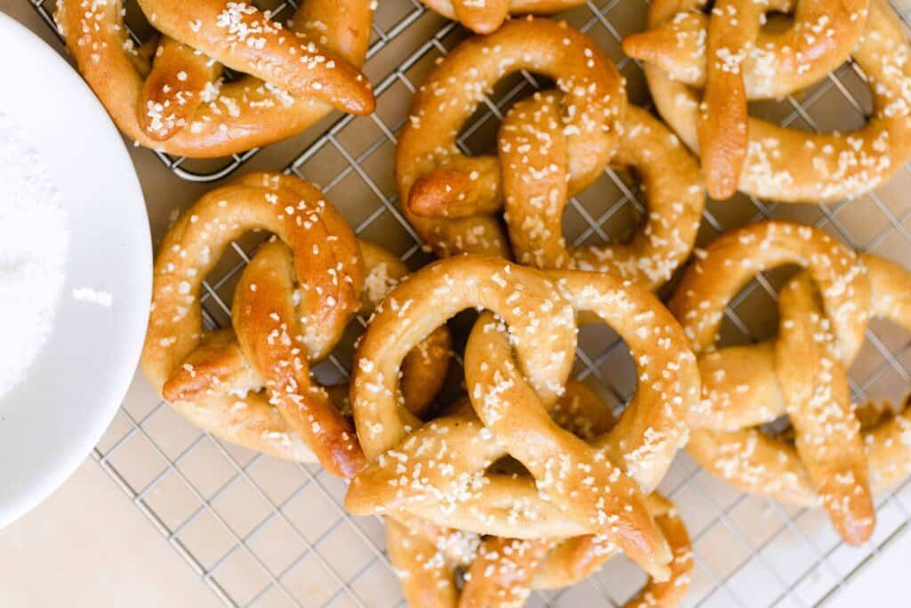 overhead photo of sourdough pretzels topped with course sea salt on a wire cooling rack with a white bowl to the left
