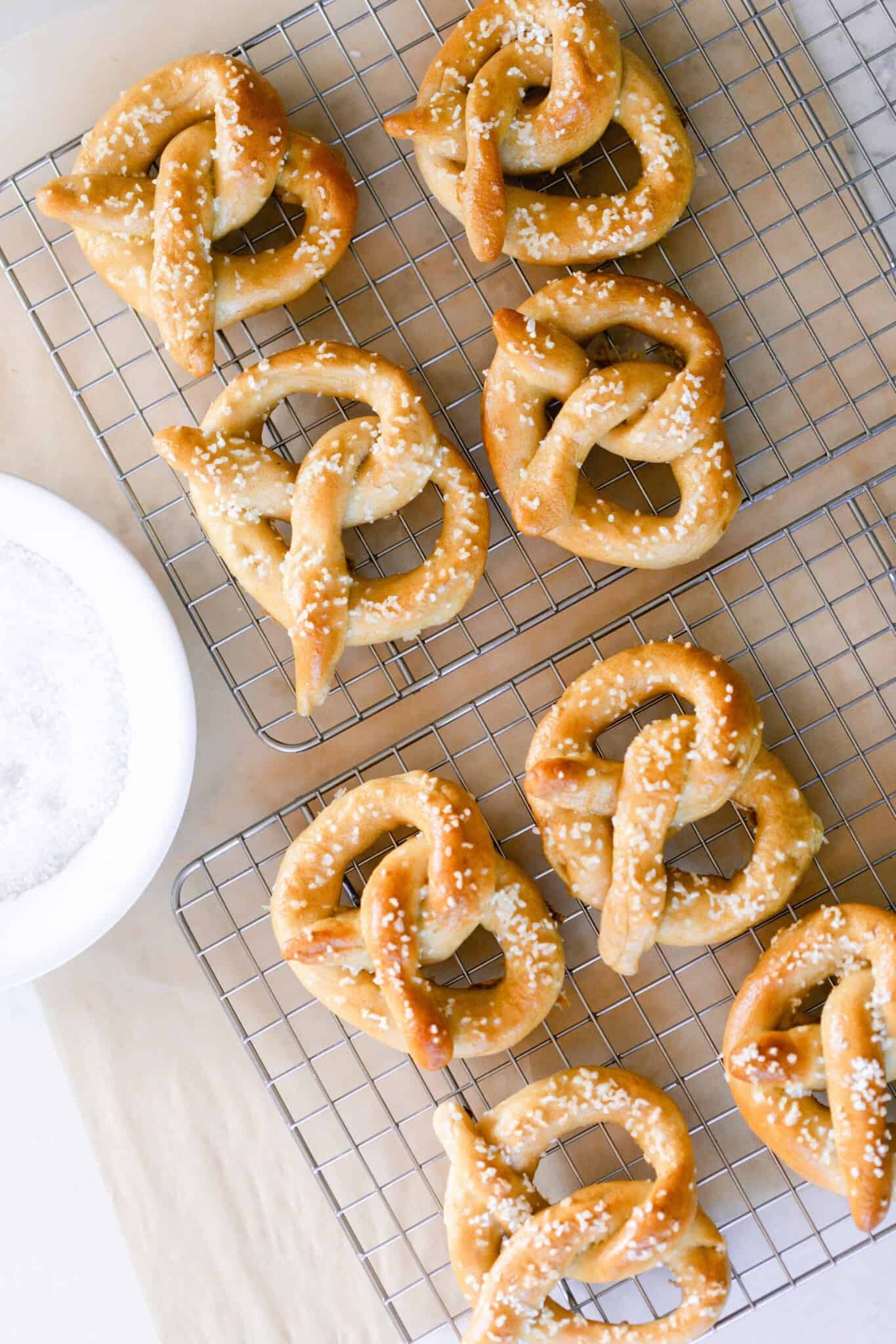 eight soft pretzels with sea salt cooling on a wire rack with a small white bowl to the left.