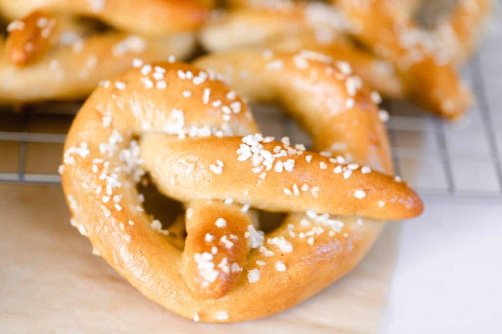 close up picture of a sourdough soft pretzel resting against a cooling rack filled with more pretzels