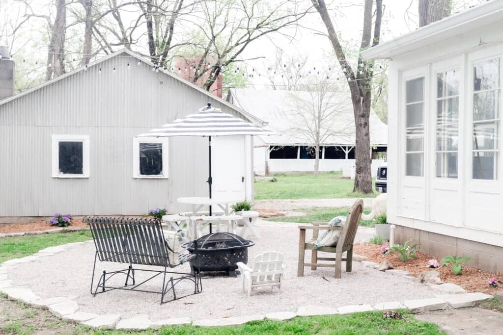 gravel patio in the back of a white farmhouse with chairs and table with an umbrella. A gray garage in the background